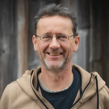 Ein Mann mittleren Alters mit Brille und beigem Kapuzenpulli lächelt vor einem hölzernen Hintergrund.