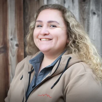 Lächelnde Frau mit lockigem blondem Haar und hellbrauner Jacke, die vor einem hölzernen Hintergrund steht.