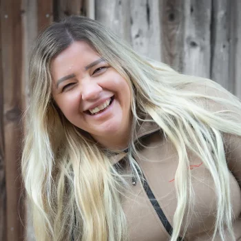 Eine lächelnde Frau mit langen blonden Haaren und einer beigen Jacke steht vor einer Holzwand.